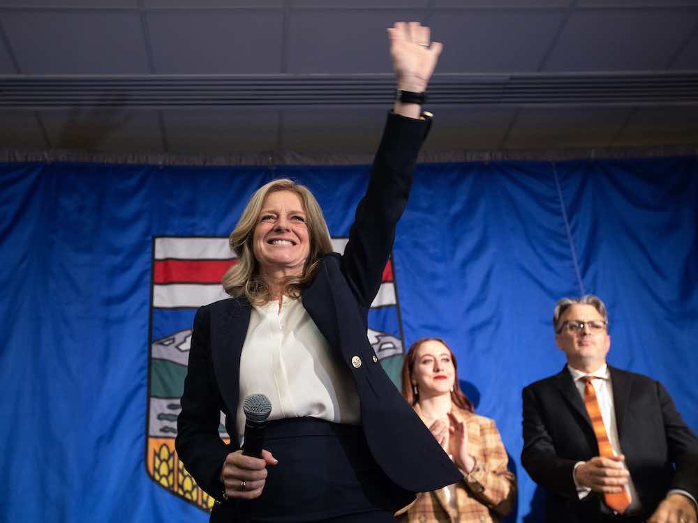 A middle-aged white woman with blond hair, wearing a dark blue jacket and white shirt, holds a microphone and waves at a crowd in a conference room.