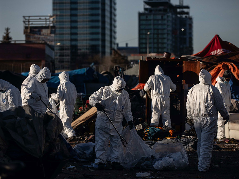 Eight people in white biohazard suits stand in front of tents. Some are loading people&rsquo;s possessions into trash bags.