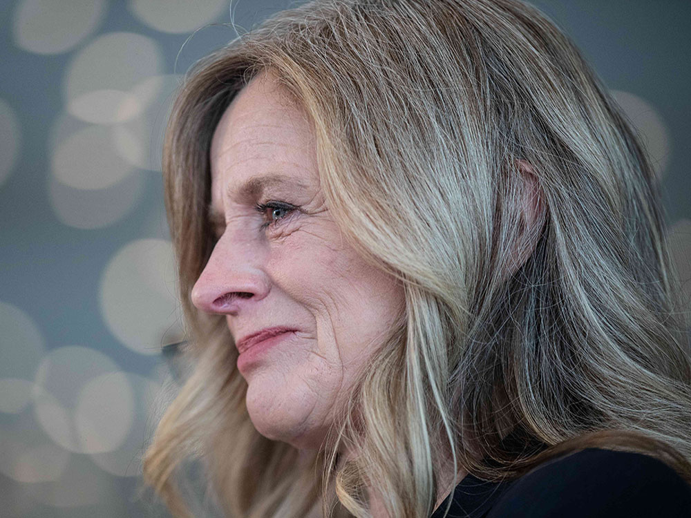 A close up of a 59-year-old white woman with blonde hair and wrinkles around her eyes and mouth, in front of an out-of-focus background.