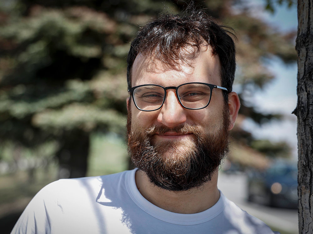 A middle-aged, light-skinned man with dark hair and a beard and large-framed glasses looks at the camera, wearing a white T-shirt. He is outdoors; the background is out of focus.