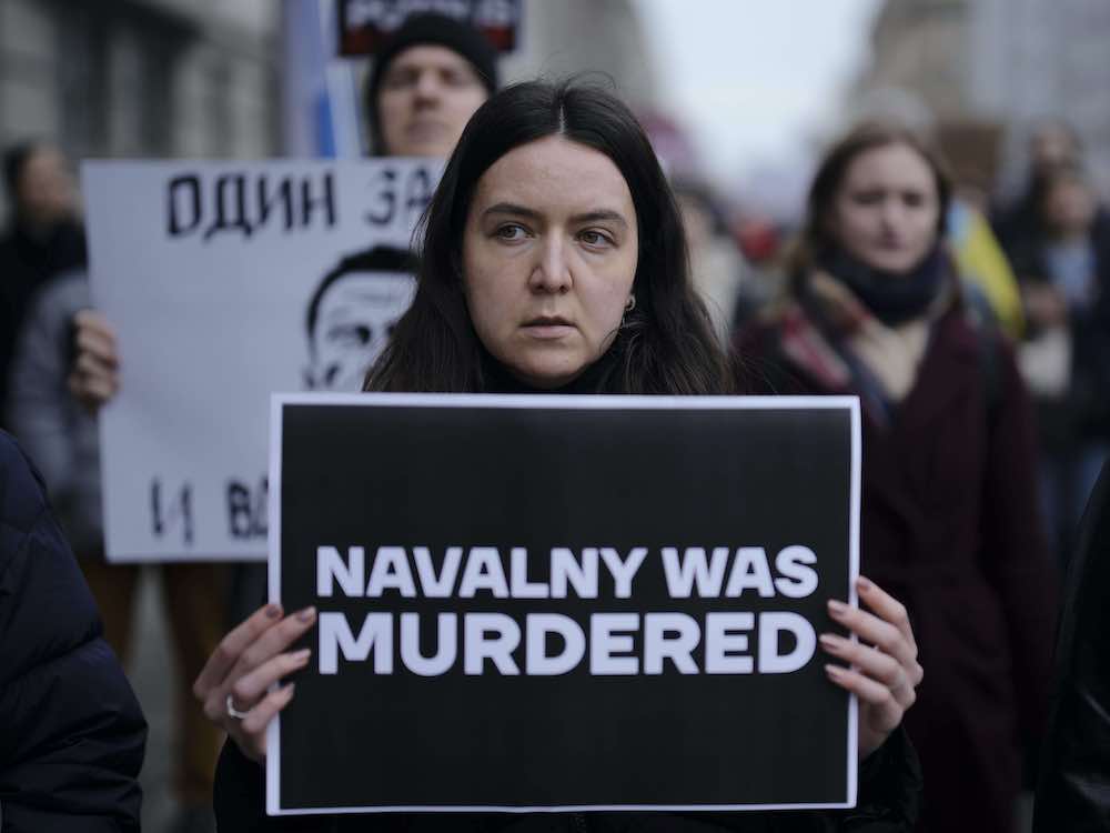 A person with long dark hair parted in the middle holds a stark black sign with white letters saying 'Navalny Was Murdered.'