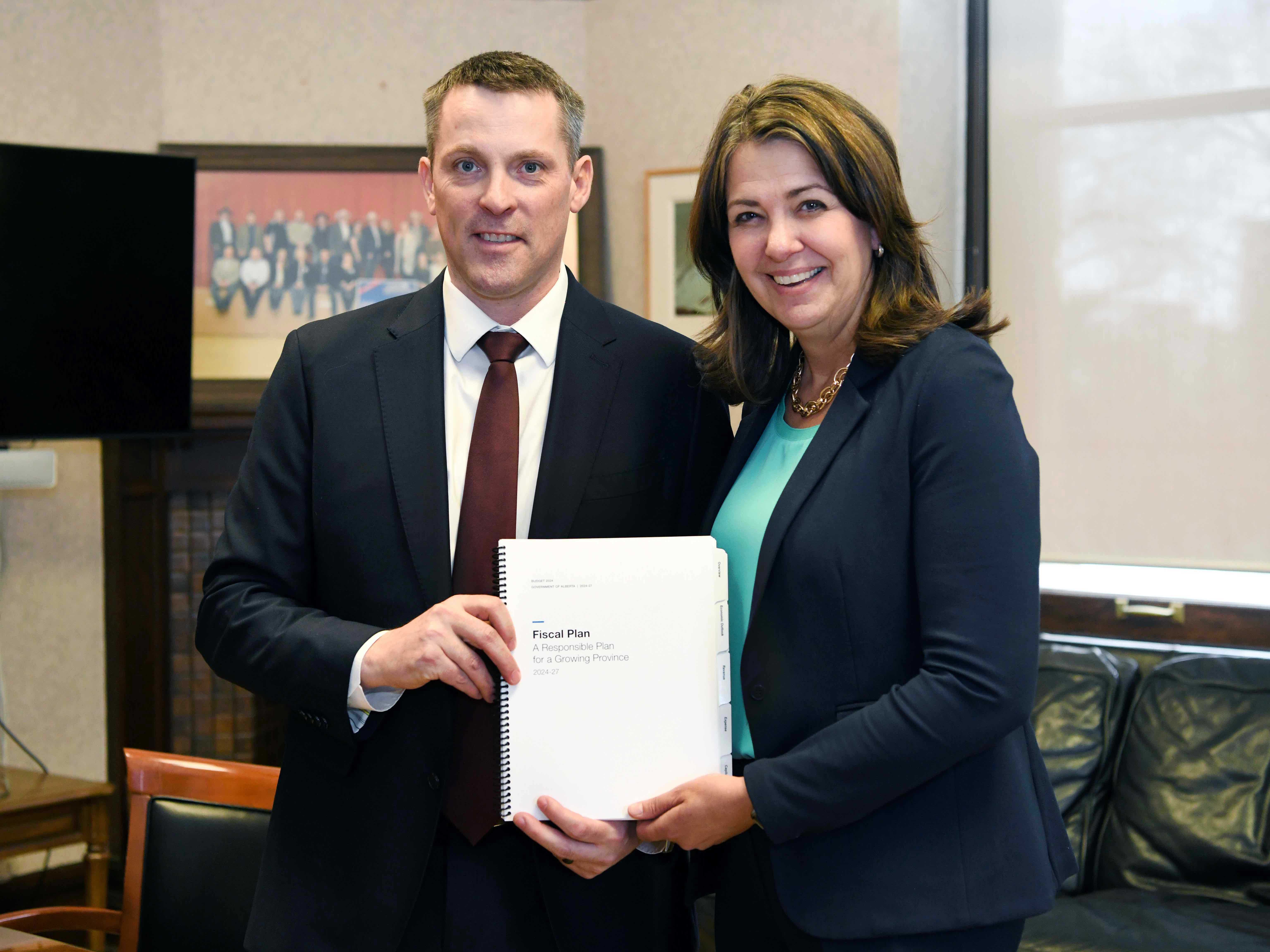 A light-skinned man with short brown hair in a dark suit and white shirt stands beside a smiling light-skinned woman with shoulder-length dark hair in a blue suit. They&rsquo;re holding a document that reads: &ldquo;Fiscal Plan: A Responsible Plan for a Growing Province.&rdquo;