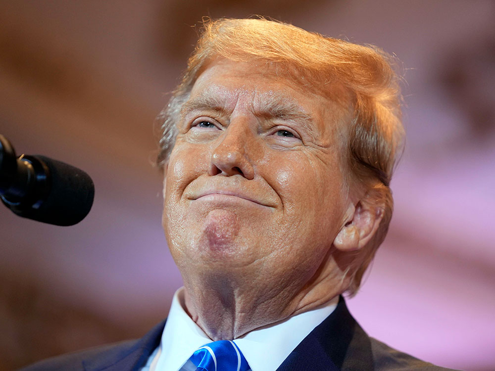 A tanned older man with styled orange hair is wearing a dark sit, white shirt and blue and white striped tie. He is offering a thin-lipped smile.