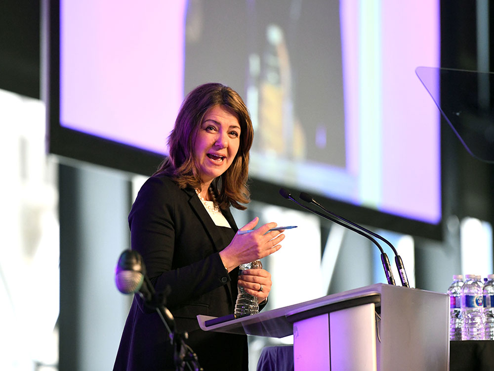 A 53-year-old light-skinned woman with shoulder-length dark brown hair stands at a podium wearing a dark suit and white shirt.