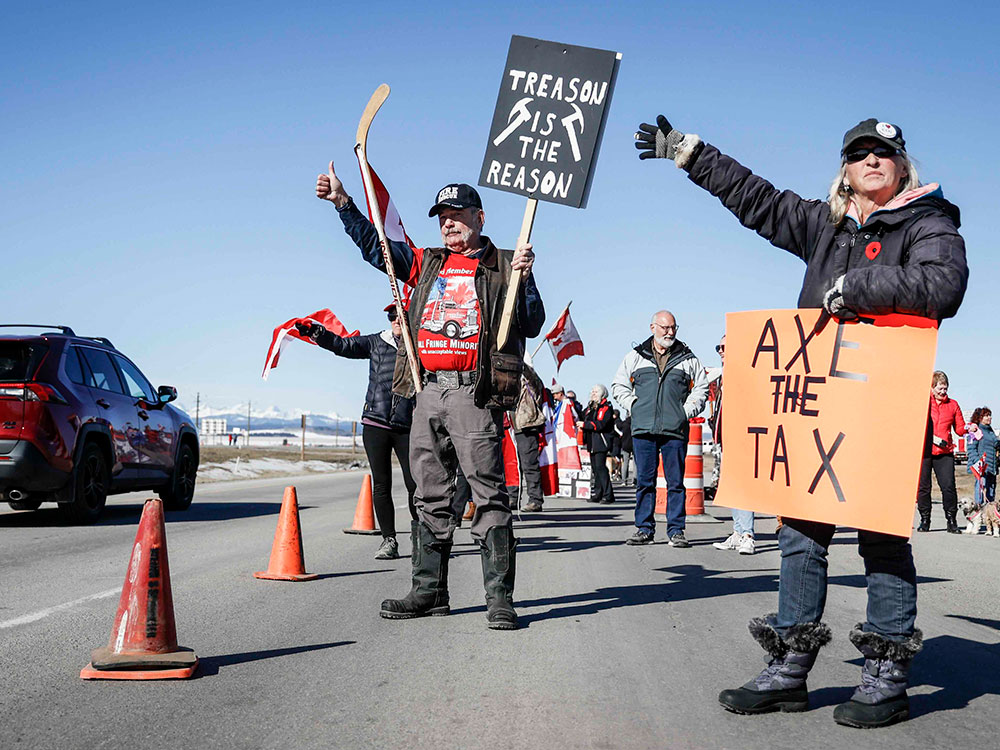 Protesters in winter jackets line a highway with signs that say 'Treason is the reason' and 'Axe the tax.' Some are wielding hockey sticks attached to Canadian flags. It’s a bright sunny day.