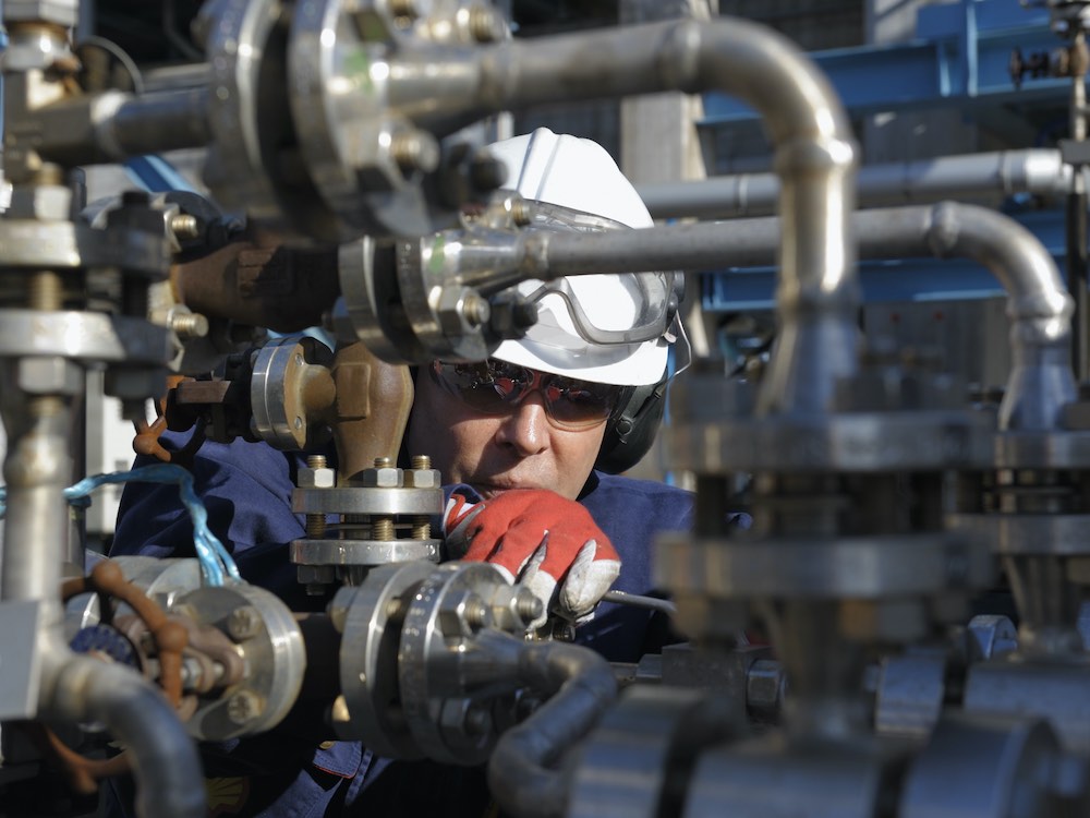 A person in a white hard hat and safety gloves peers through a tangle of silver pipes and valves.