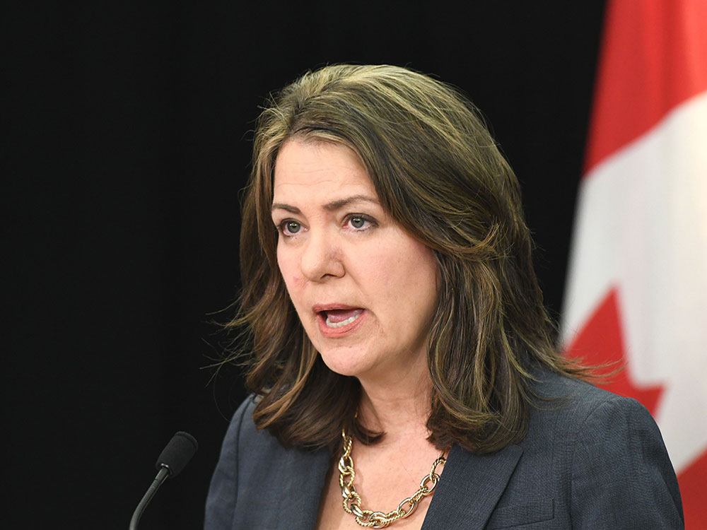 A middle-aged woman with streaked brown hair, wearing a grey jacket and gold necklace, speaks at a podium.