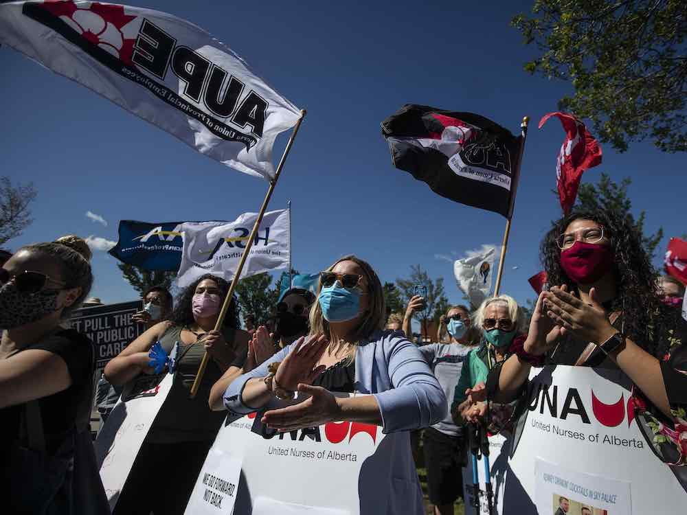 Protesters stand under a deep blue summer sky, holding flags and signs that say 'United Nurses of Alberta.'