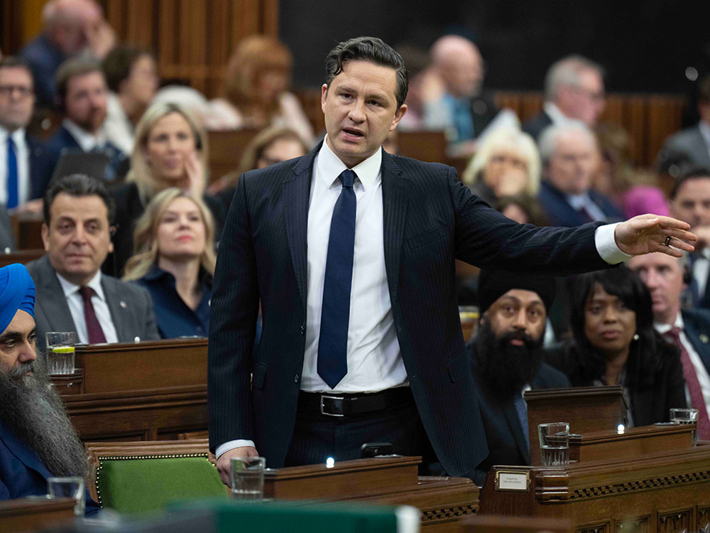A 44-year-old white man with short black hair gestures, standing in front of rows of MPs. He wears a dark suit, crisp white shirt and dark tie.