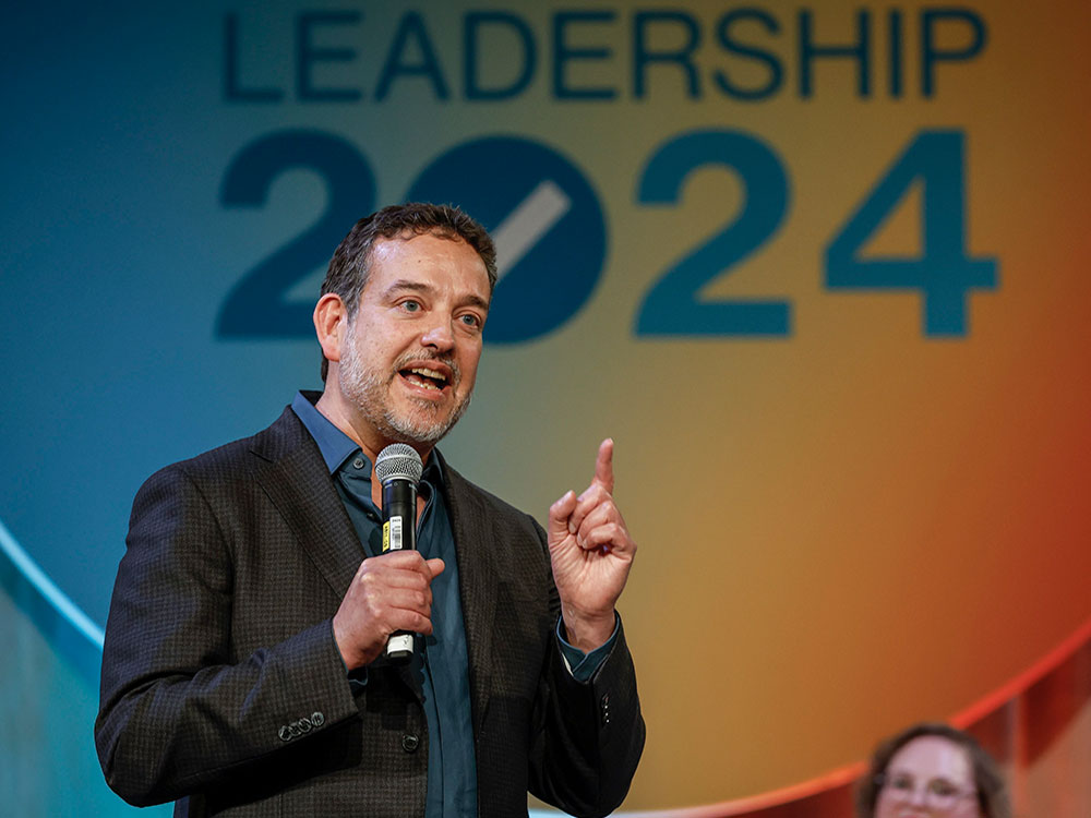 A light-skinned man in his mid-50s speaks on a stage with ‘Leadership 2024 on a screen behind him. He has short dark hair and a trimmed greying goatee, and wears a blue shirt and dark jacket.
