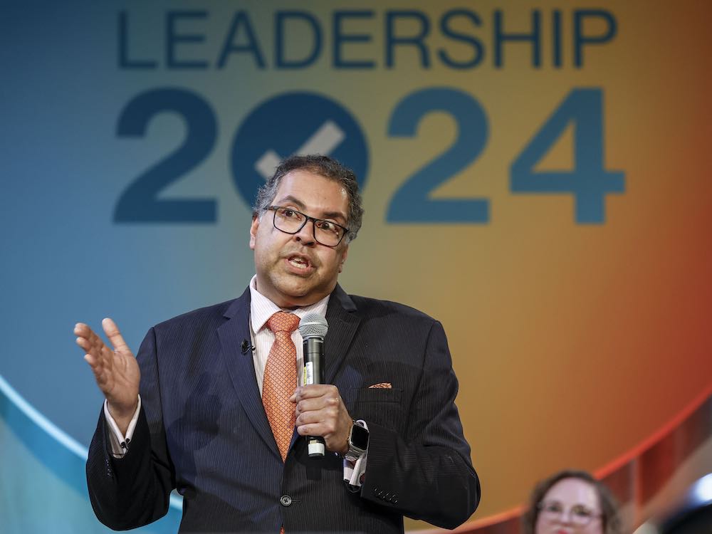 A 52-year-old man with medium skin tone holds a microphone and stands on a stage, with the words 'Leadership 2024' projected behind him. He wears a dark suit, white shirt and orange tie and gestures.