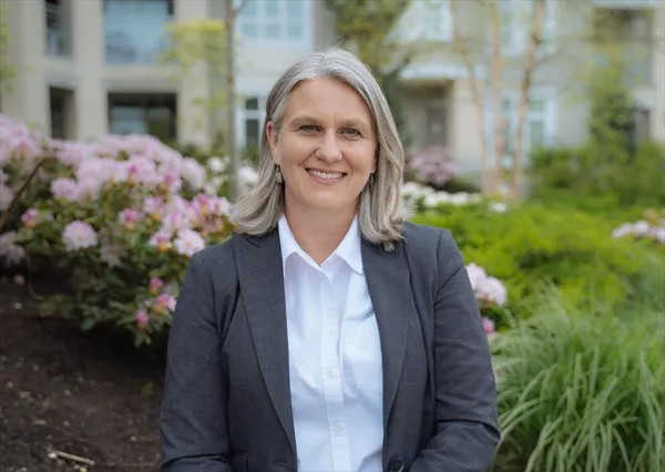 A middle-aged person with light skin and blond hair smiles at the camera. She stands in a garden and wears a white shirt and grey jacket.
