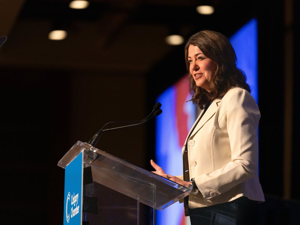 A 53-year-old light-skinned woman with shoulder-length brown hair stands smiling at a podium. She wears a stylish white jacket.