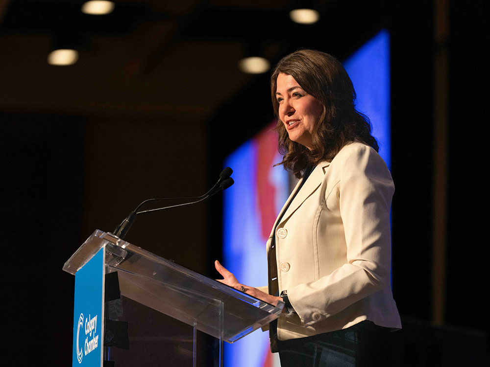 A middle-aged woman with wavy, shoulder-length brown hair and a beige blazer stands at a podium on a darkened stage. She is smiling and looking to the right of the frame.