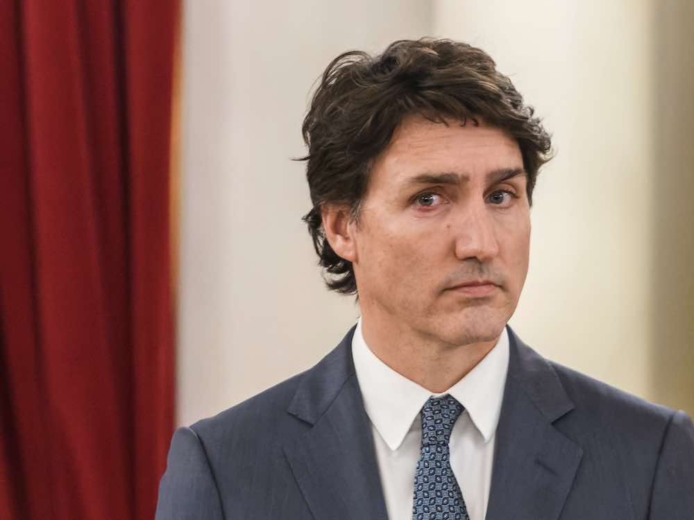 A light-skinned 52-year-old man with styled hair and a light grey suit and white shirt looks at the camera without smiling. He appears tired.