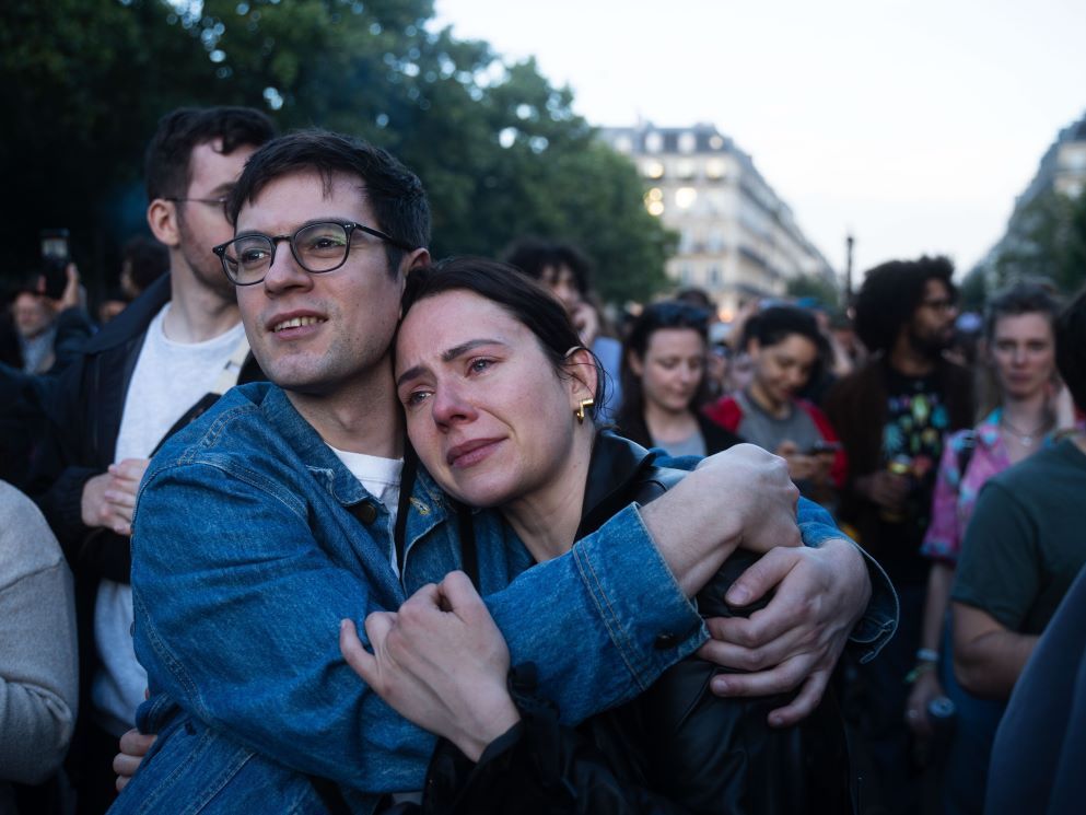 A man and woman with light skin embrace while walking in a crowd of people. He is smiling; she is slightly smiling and looks as if she has been crying.