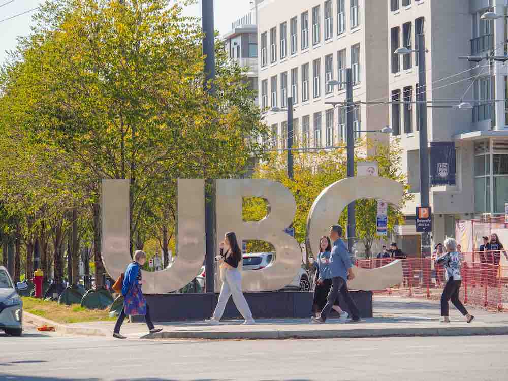 People walk on the sidewalk past a large silver outdoor installation of the letters 'UBC' on a sunny day.