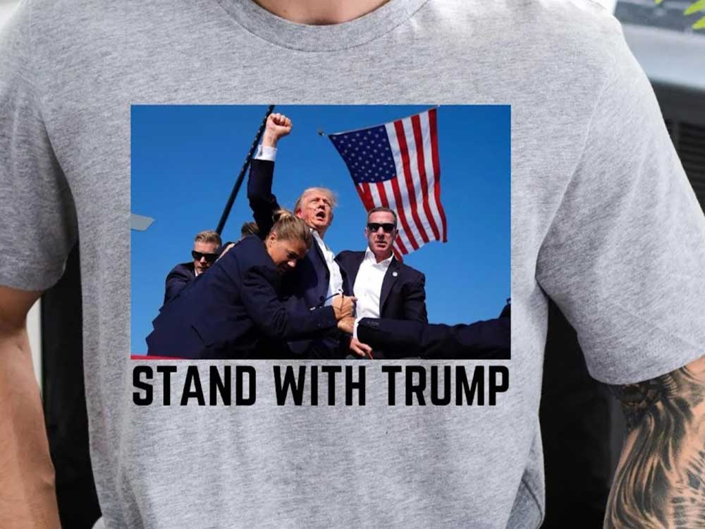 A T-shirt has a photo printed on it showing Donald Trump raising his fist triumphantly, with Secret Service agents around an American flag in the background.