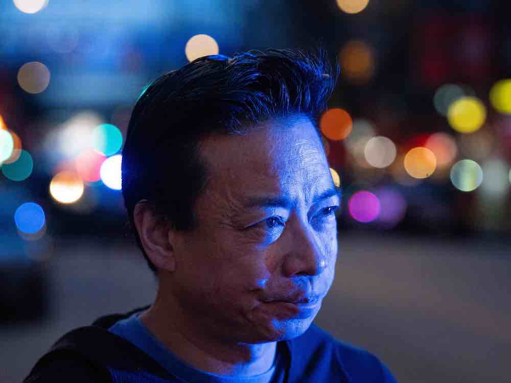 A 53-year-old man with styled dark hair looks intent. It&rsquo;s night and he&rsquo;s illuminated by blue lights.