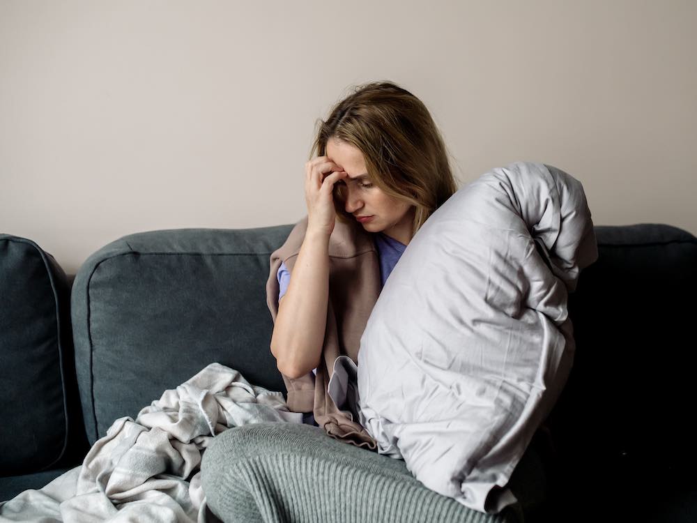 A woman sits on a couch, clutching a pillow and looking tired, with a blanket beside her.