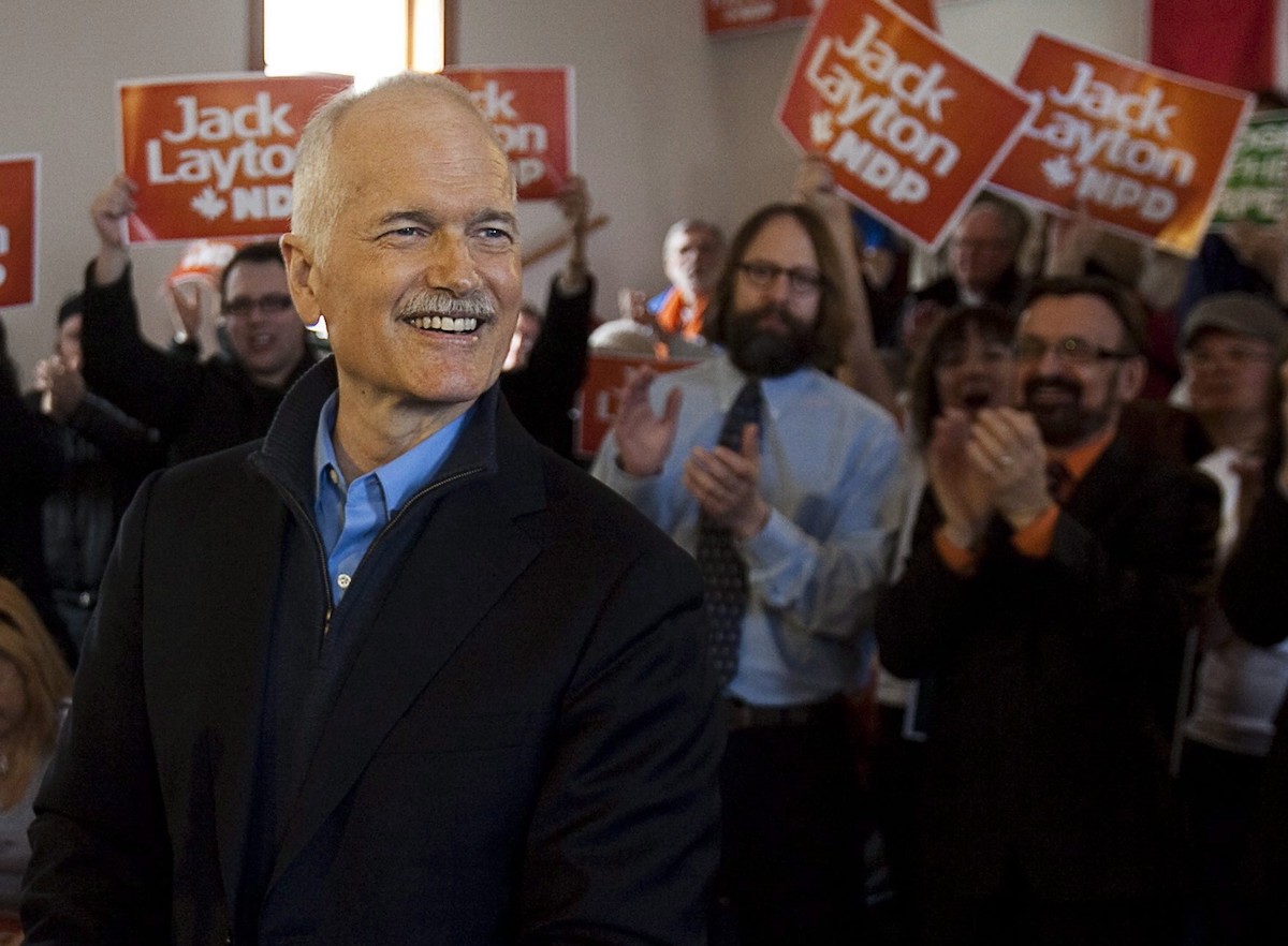 A white middle-aged man, Jack Layton, in a white shirt with sleeves rolled up, smiles. In the background are men and women with signs supporting him as NDP leader.