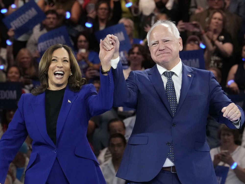 Kamala Harris, left, wears a blue blazer over a black top. She has medium skin and shoulder-length brown wavy hair. Her left arm is raised to clasp the hand of Tim Walz, right, a man with white hair, light skin and a navy suit over a white shirt. They’re both smiling, and a crowd holds blue signs behind them.