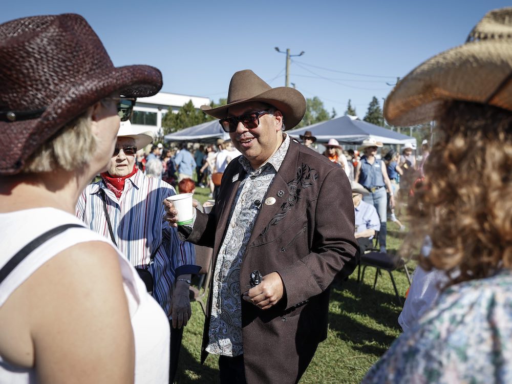 A middle-aged man with medium skin tone stands outside chatting with people. He wears a brown jacket and cowboy hat.