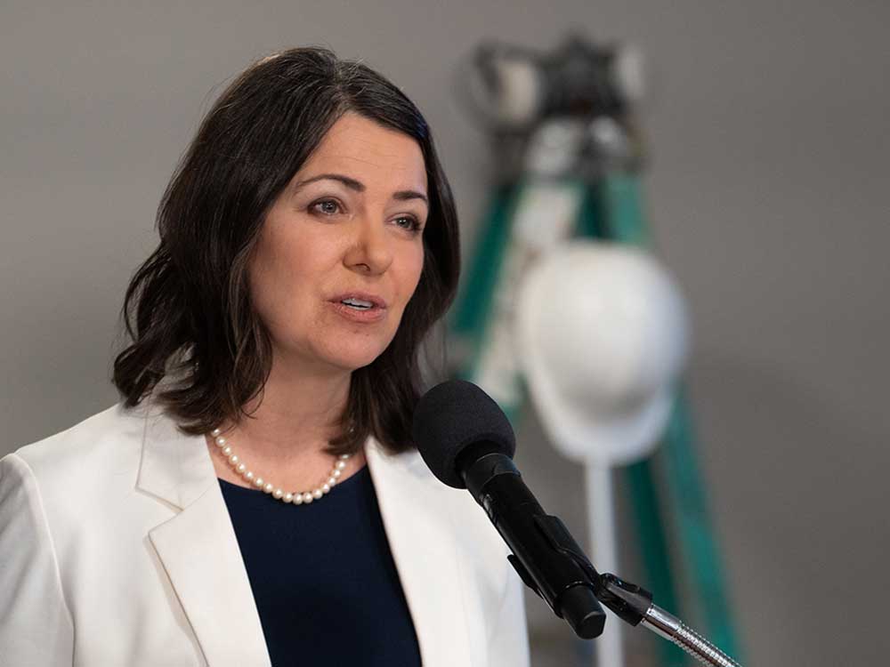 A 50-ish light-skinned woman with shoulder-length brown hair stands at a microphone. She wears a white jacket.