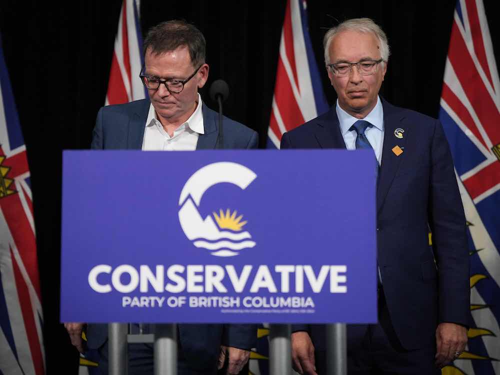 Two light-skinned older men wearing suits stand behind a blue sign that says 'Conservative Party of British Columbia.'
