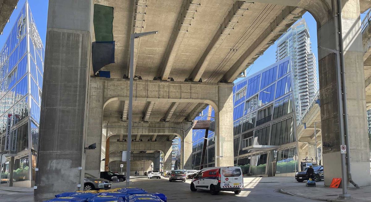 The underside of a large, concrete bridge. Glass towers are visible from the openings on either side, and cars drive and are parked below.