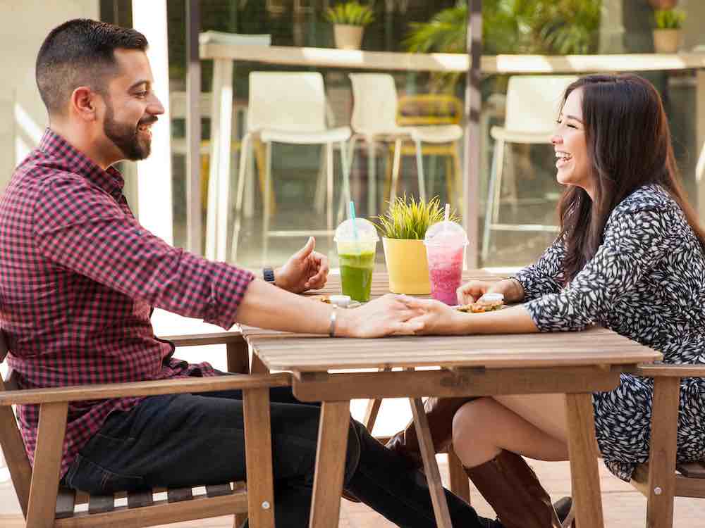 Two people hold hands across a wooden table on an outdoor patio at a café. Two iced beverages in green and pink are on the table. On the left, a man with short dark hair, a beard, medium skin and a buffalo plaid shirt is smiling at the woman, right, across from him. The woman has long dark hair, light skin, and wears a blue and white floral dress.