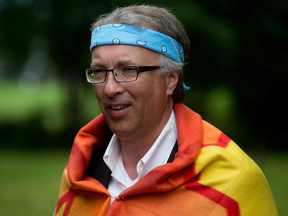 John Rustad is a white man with short grey hair and glasses. He is standing in an outdoor setting wearing a light blue bandana as a headband and a yellow and orange blanket as part of a Sto:lo First Nation blanketing ceremony. The blanket is fastened with a safety pin over Rustad’s white button-down shirt.