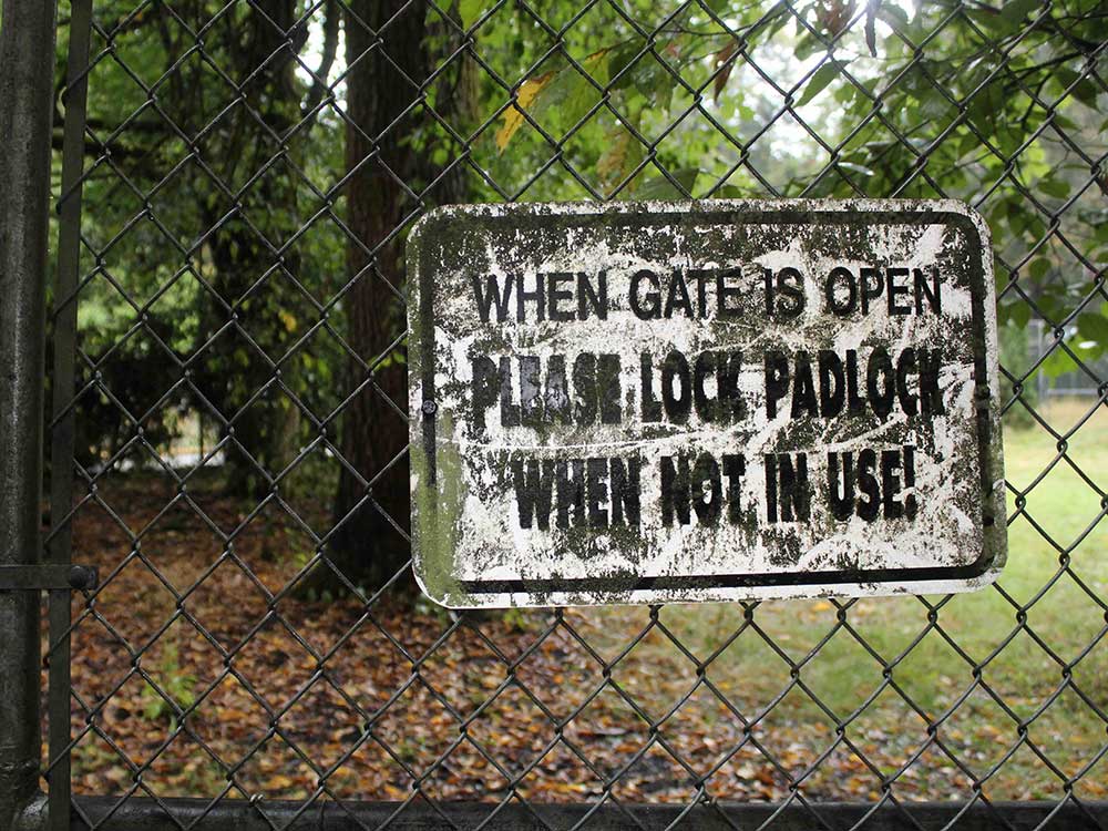 A black chain-link fence carries a worn white sign that reads, in black text, &ldquo;When gate is open please lock padlock when not in use!&rdquo; In the background is a shaded grassy area with trees.