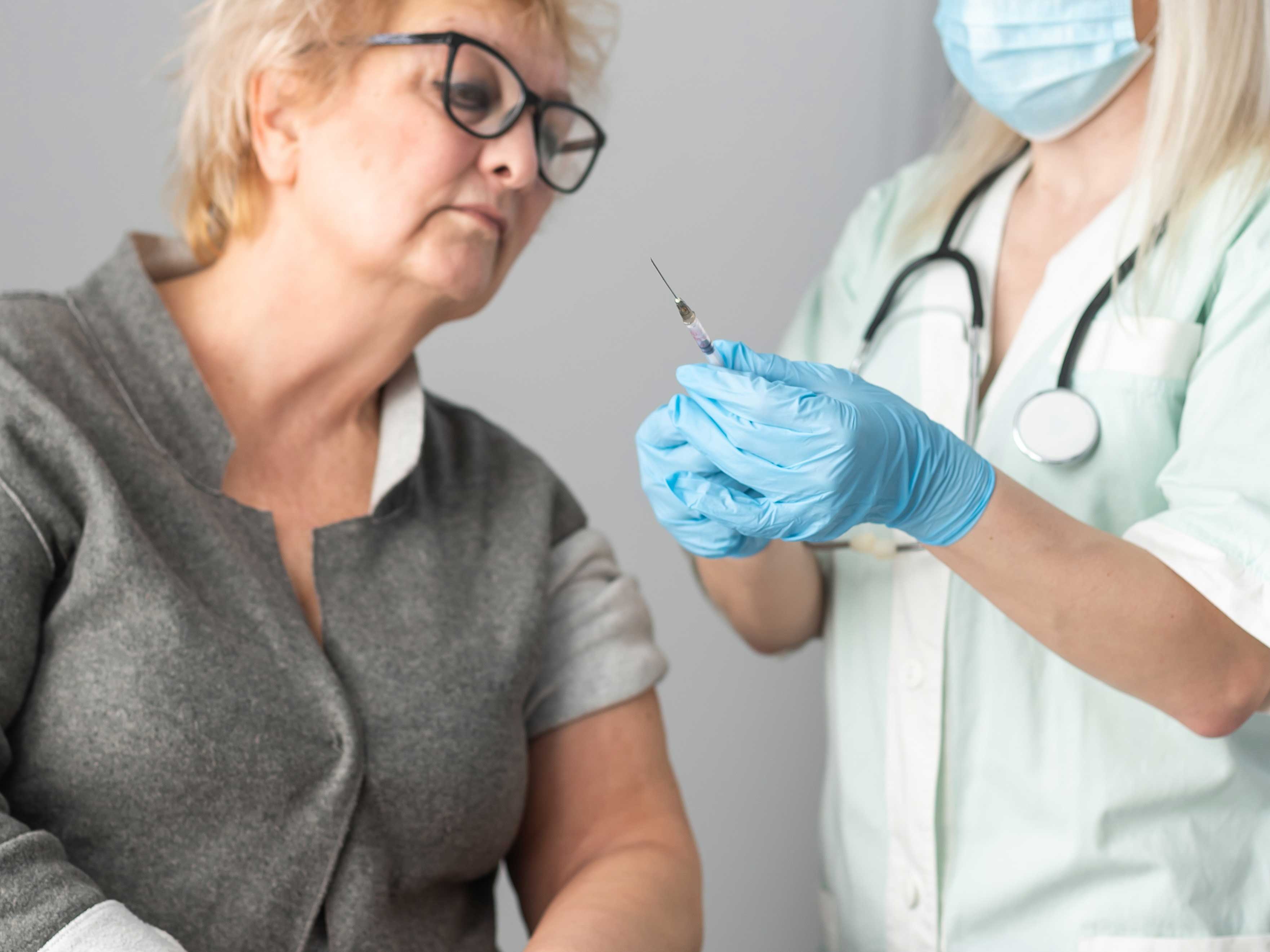 An older light-skinned woman looks at a syringe held by a light-skinned woman wearing medical garb.