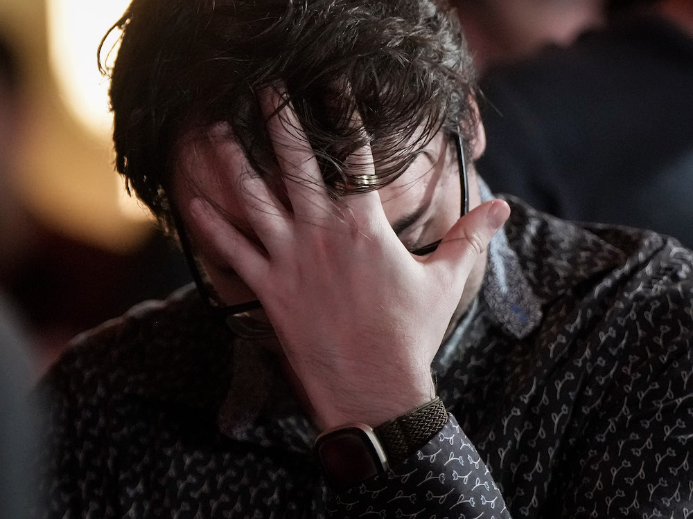 A young man with tousled brown hair anxiously holds his hand over his face.