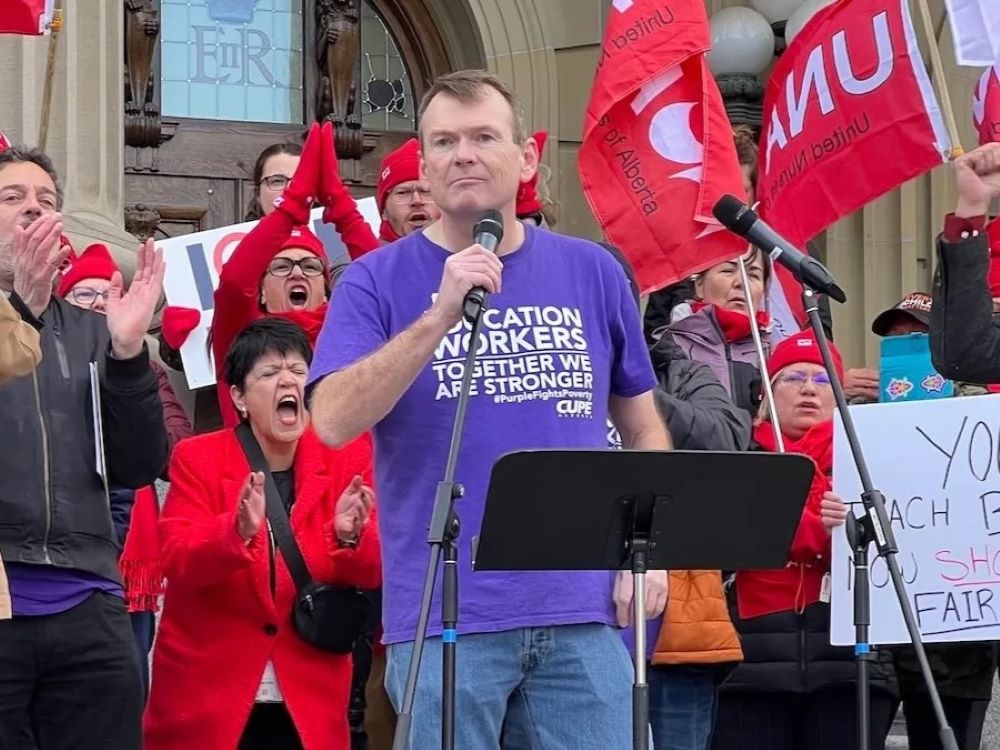 A 40-ish light-skinned man in a purple T-shirt holds a microphone. He stands in front of an enthusiastic group of people, some holding red flags.