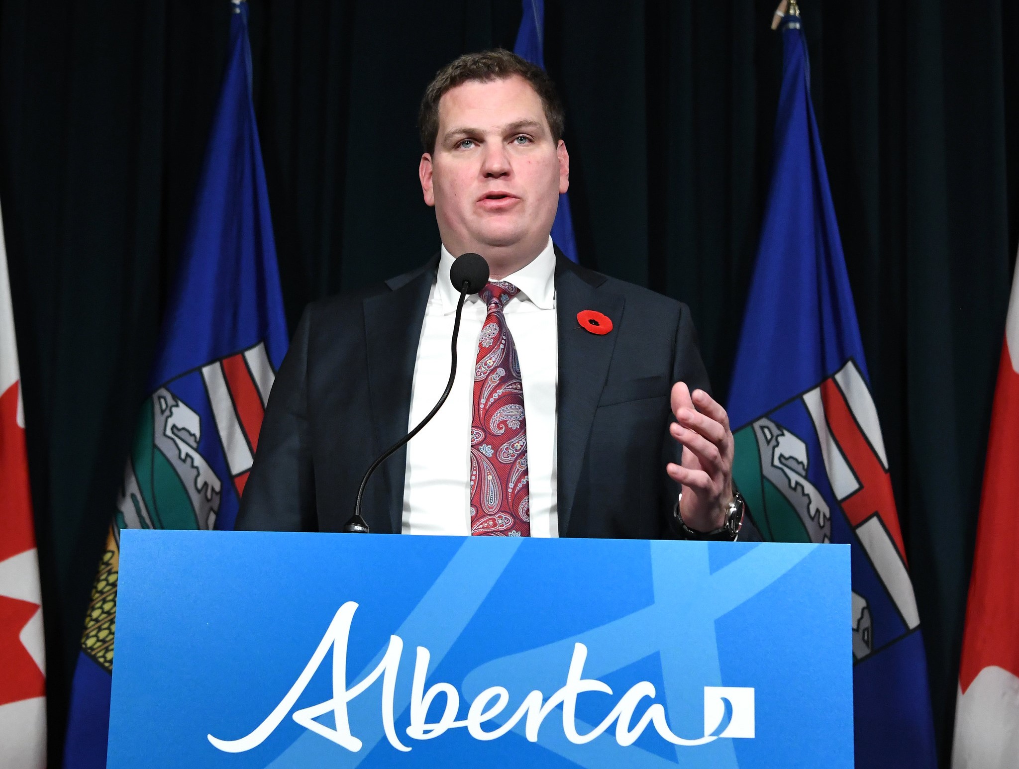 A middle-aged light-skinned man wearing a dark suit, white shirt and tie stands at a podium.