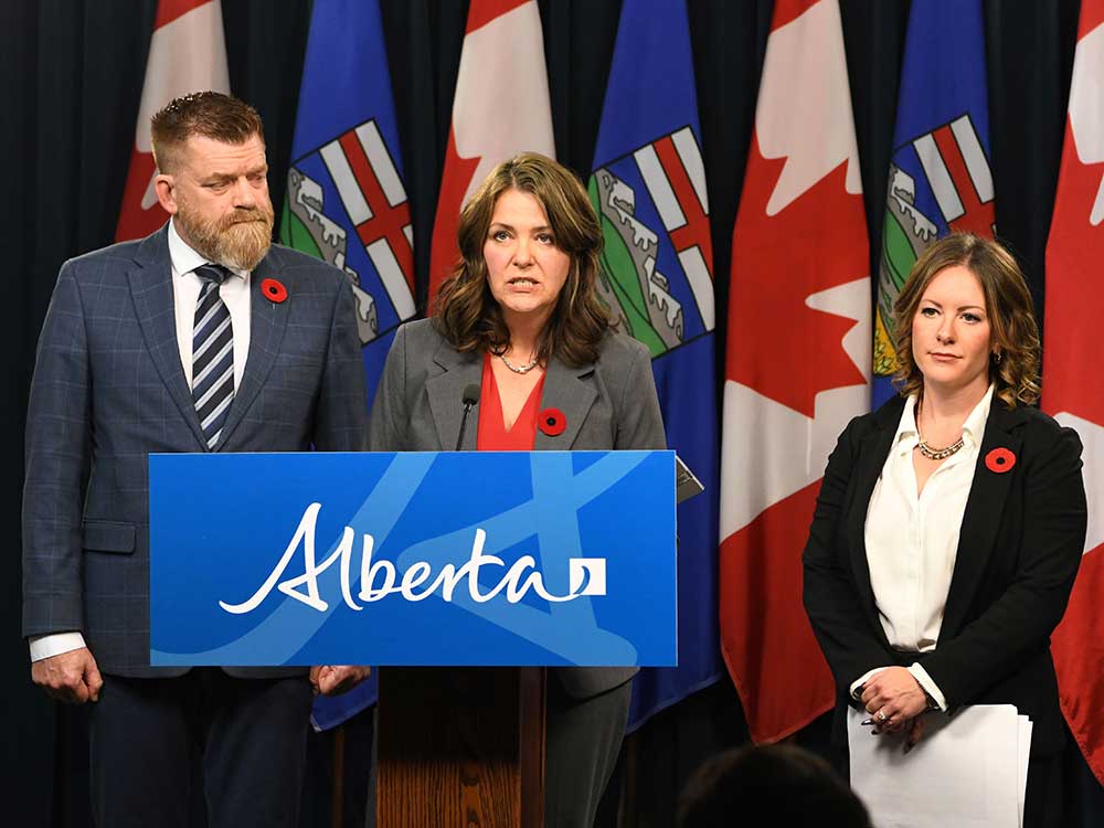 A 60-ish woman with light-toned skin and shoulder length dark hair stands at a podium in front of flags. A 61-year-old light skinned man with a large beard stands at her left, a 40-year-old light-skinned woman at her left.