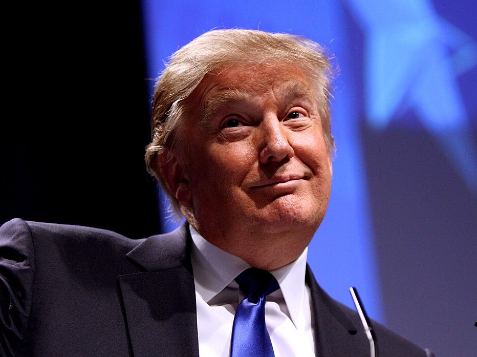 A closeup of Donald Trump smiling with his mouth closed, raising his eyebrows and looking upward. He wears a suit and blue tie. 