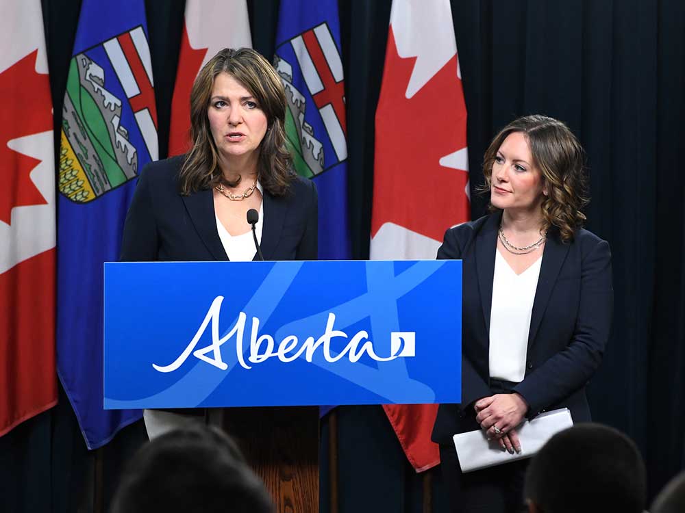 Two light-skinned women with shoulder-length brown hair stand in front of flags at a media event. They wear blue suits, white shirts and necklaces.