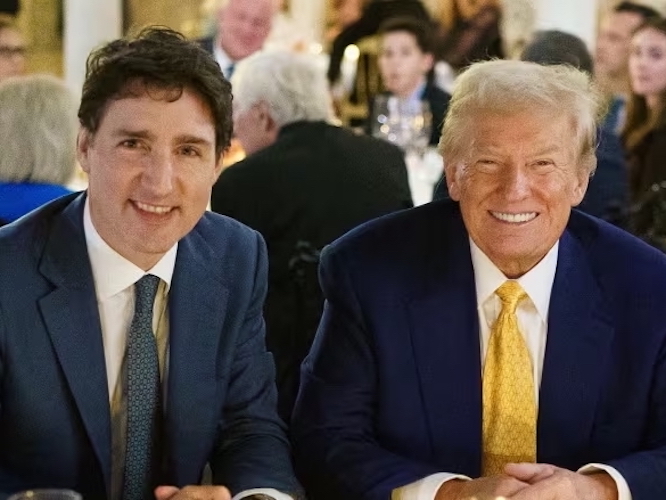 A 52-year-old light-skinned man sits at a dinner table beside a 78-year-old man with an orangish complexion. Both wear suits and smile at the camera.
