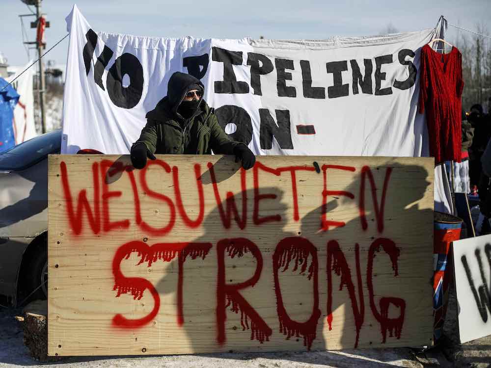 A person wearing heavy winter clothes stands behind a roughly painted sign that says 'Wet&rsquo;suwet&rsquo;en strong.' Behind is a sign that says 'No pipelines.'