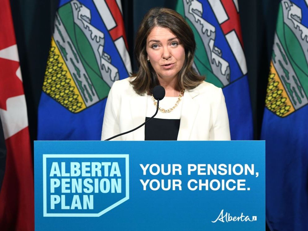 Danielle Smith, a light-skinned woman in her mid-50s with shoulder-length brown hair, wears a white blazer and gold necklace. She stands at a podium with a sign that says &lsquo;Alberta Pension Plan, Your Pension, Your Choice.&rsquo;