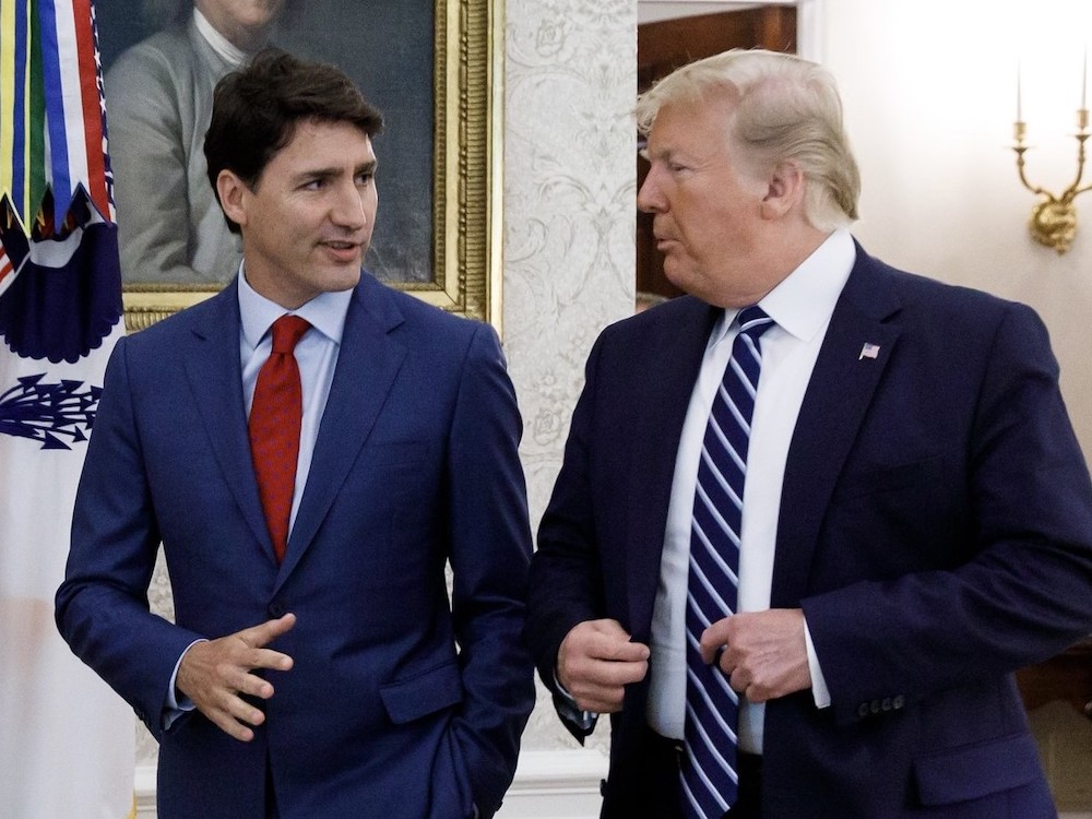 Justin Trudeau and Donald Trump converse while walking across an indoor space behind a white sofa and in front of a set of flags and an ornate wall hanging. Trudeau has short dark hair and is wearing a dark blue suit over a light blue shirt and red tie. Trump has wavy blond hair and is wearing a navy suit over a white shirt and a diagonally striped blue and white tie.