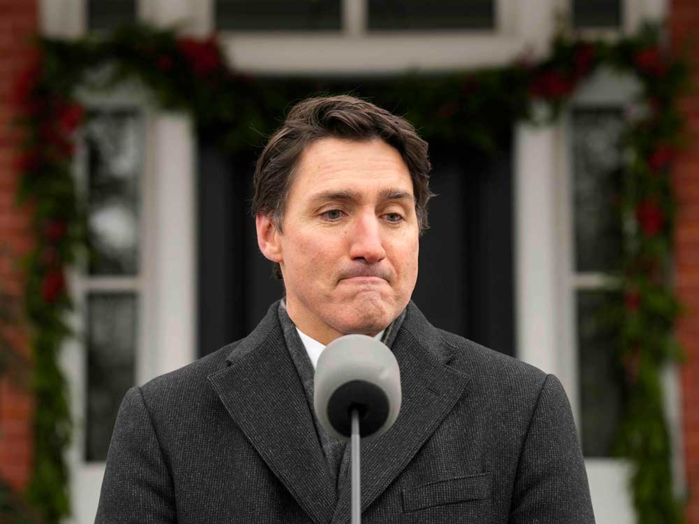 Justin Trudeau, a 53-year-old light-skinned man, stands outdoors at a podium wearing a dark overcoat. He looks discouraged.