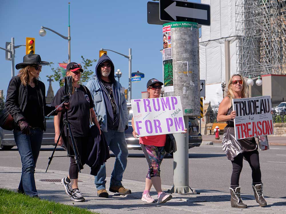 A group of people walk on the sidewalk outside the Parliament Buildings. Two women carry signs, one saying “Trump for PM” and the other saying “Trudeau for treason.”