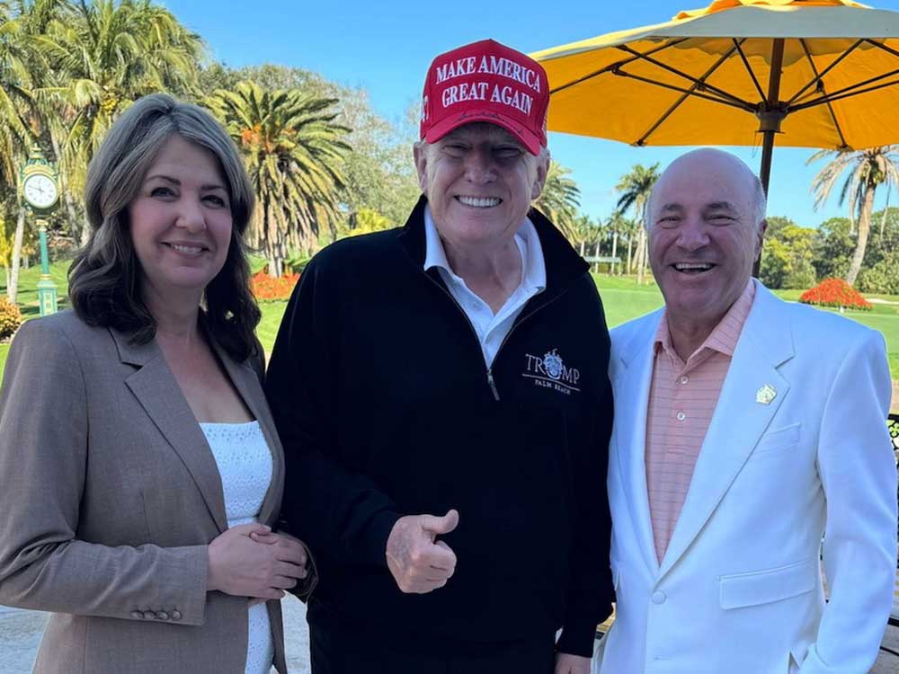 Three older light-skinned people smile at the camera on a golf course. Danielle Smith wears a brown suit and white shirt. Donald Trump wears a dark pullover and red MAGA hat. Kevin O’Leary wears a white suit.