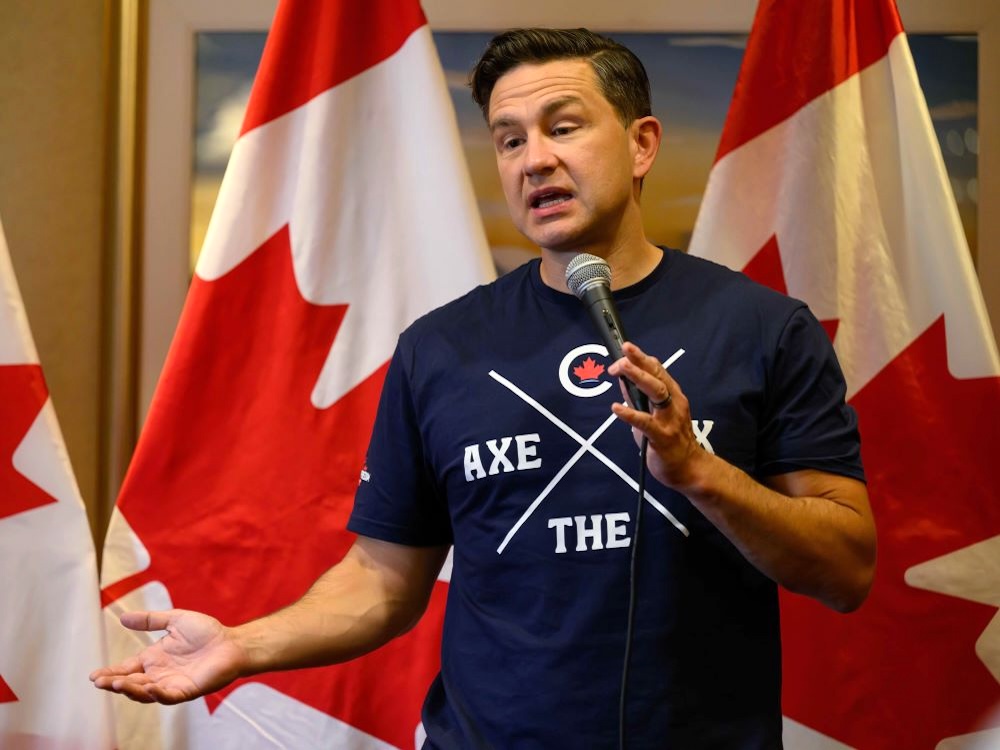 A 45-year-old light-skinned man holds a microphone while standing in front of Canadian flags. He wears a dark T-shirt that says 'Axe the Tax.'