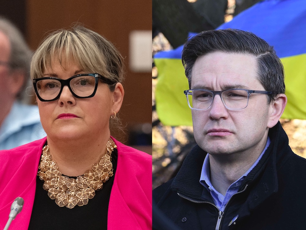 On the left, a middle-aged woman with light skin, streaked blond hair and black-framed glasses wearing a magenta jacket over a black top and an ornate necklace made up of gold flowers. On the right, a middle-aged man with light skin, dark hair and square-framed glasses wearing a black zip-up jacket. Both are unsmiling and slightly to the left.