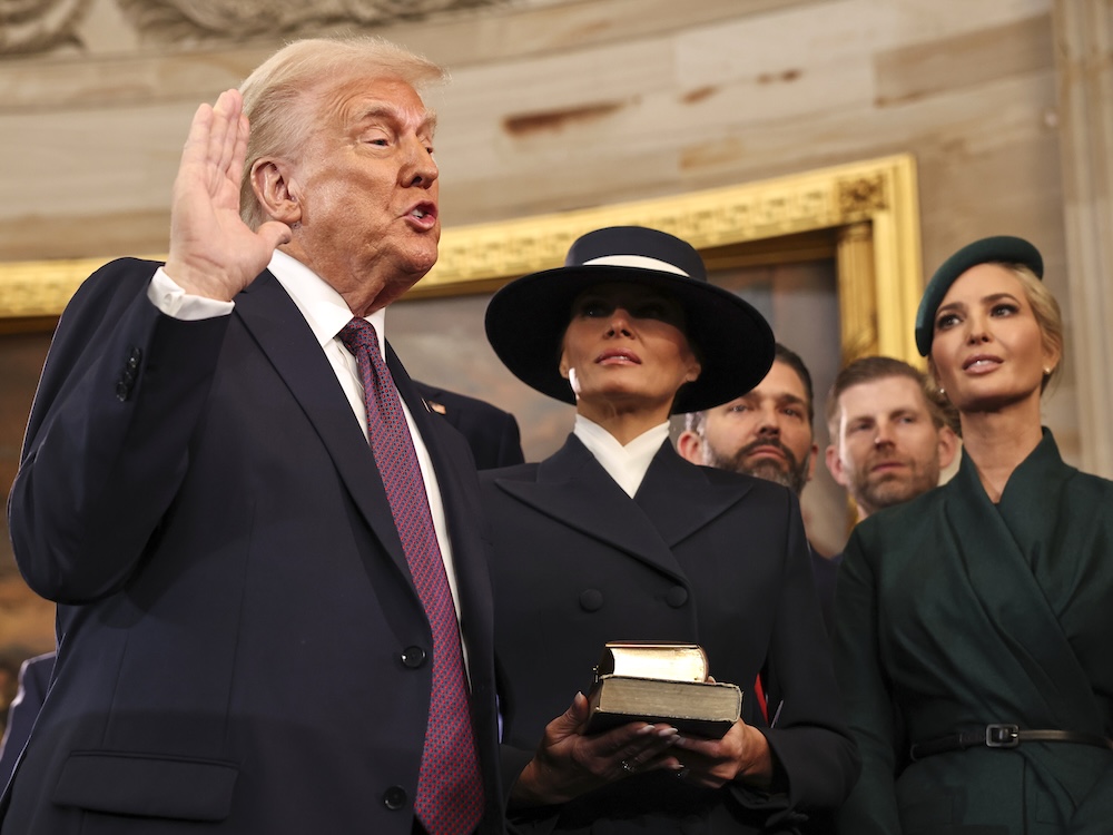 A 78-year-old man in a suit with coiffed blondish hair holds up his right hand. A woman in a dark suit and wide-brimmed hat looks on, holding two books. So does a younger woman with blond hair wearing a green suit and matching hat.